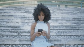Pretty young Afro-american woman with curly hair and in jeans sleeveless dress sitting outside, googling on phone, looking surprised, saluting. Lifestyle, communication concept