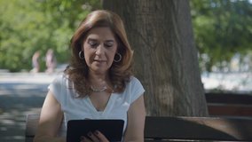 Medium shot of elegant middle-aged woman in white blouse sitting on bench outside, googling on tablet, nodding head, being surprised, then satisfied. Lifestyle, modern technology concept