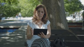 Medium shot of elegant middle-aged woman in white blouse sitting on bench outside, googling on tablet, talking. Lifestyle, modern technology concept