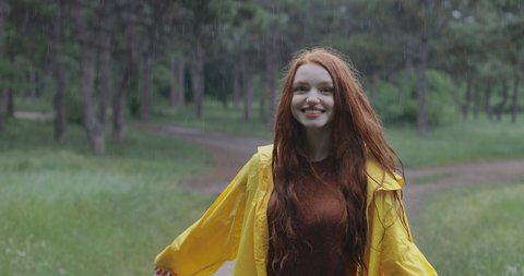 Attractive red-haired young woman in a yellow rain coat walking in the forest. The girl laughs and rejoices in the spring warm rain. tracking shot. in slow motion. Shot on Canon 1DX mark2 4K camera