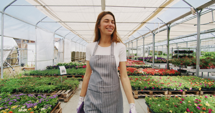 Slow motion of an young woman gardener in apron is walking satisfied with her work in a plant shop greenhouse in a sunny day. Royalty-Free Stock Footage #1030525136