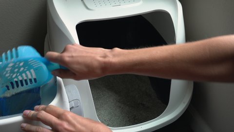 Woman cleaning a cat litter box by scooping litter clumps with a blue scoop into a trash disposal box.