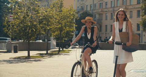 Two women walking together on street. Lifestyle and health in city. Girls riding vintage bike and electric scooter on urban background. 4K slow motion video footage 60 fps