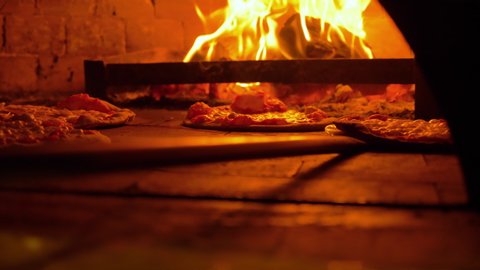 Restaurant chef Italian pizza is cooked takes pizza  in a wood fired oven at traditional restaurant.Close up pizza in firewood oven with flame behind being pulled from mobile wood fired oven 