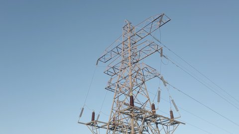 Low angle tracking shot of high voltage electrical supply pylon tower set against blue sky.