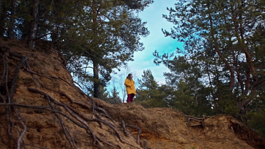 Woman in a yellow jacket on top of a ravine in the forest, she photographs a beautiful blue sky with clouds, spring travel, nature.