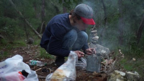 A man prepares food at the stake.The guy in the cap kindles a fire in the woods. Picnic in the woods. Metal mug with coffee on the fire. Dinner at camp. Food during the journey.

