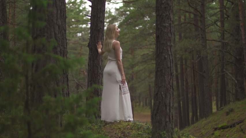 Walking Beautiful young blonde woman forest nymph in white dress in evergreen wood
