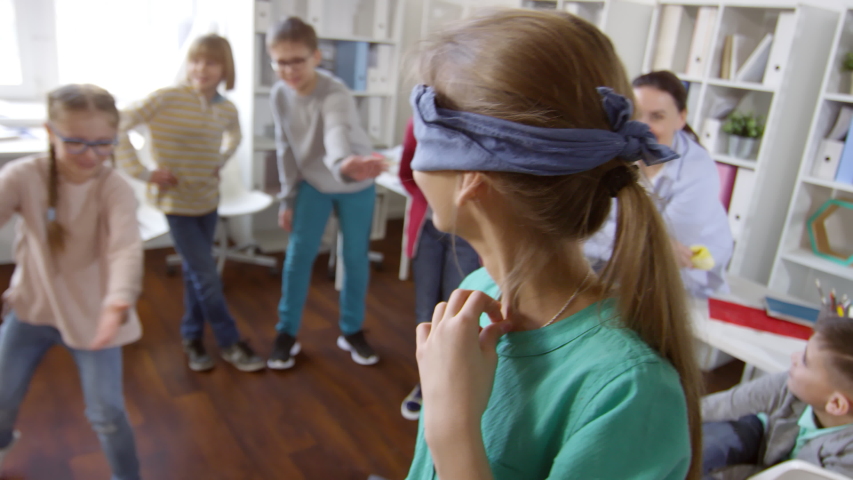 Waist-up shot of laughing girl wearing blindfold listening to her friend’s directions, then slowly stumbling around cushions with outstretched arms, while other kids watch them in background