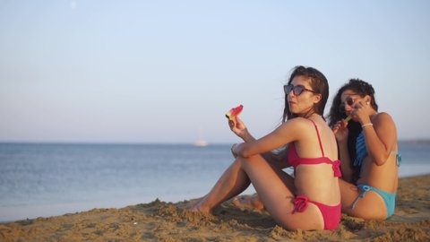 Happy hipster teenage girls on summer vacations sitting on sandy island beach and eating watermelon
