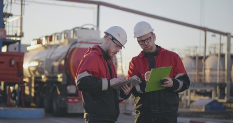 Industrial male professional engineers at oil and gas refinery use tablet and discuss data from working documents. 16 wheeler tanker on the background. Industrial chemical business concept