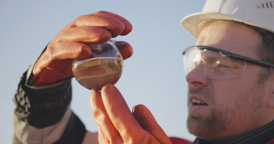 Close portrait of handsome experienced male caucasian bearded engineer in uniform and hardhat examining oil solution or substance in test tube. Refinery, exploration, oil and gas industry