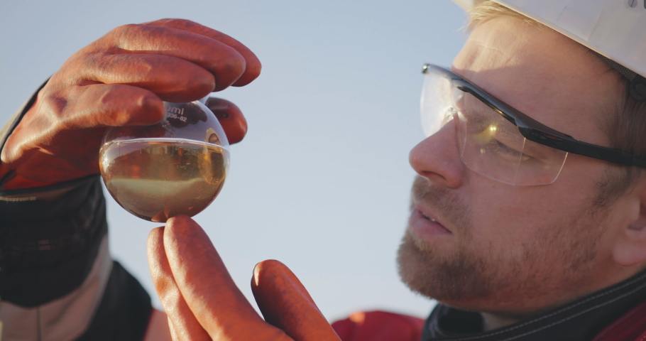 Close portrait of handsome experienced male caucasian bearded engineer in uniform and hardhat examining oil solution or substance in test tube. Refinery, exploration, oil and gas industry