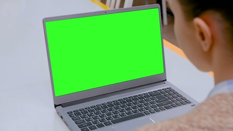 Woman sitting at white table and looking at grey laptop computer device with blank green screen in cafe, home or office. Technology, chroma key, template, mockup and entertainment concept