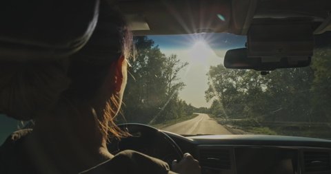 Woman drives the car while sunset.  Young woman driving the car.  Young adult girl driving car. Real time. Sun's rays shine through the windshield of the car. Rear View to woman drives a automobile 