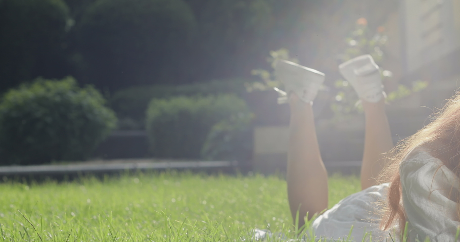 Cute red-haired girl smiling lying on the grass near the house. Young woman wiggles her legs and looks happy and fun. tracking shot. real time. Shot on Canon 1DX mark2 4K camera