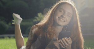 Beautiful girl in headphones with red hair and freckles listens to music using a smartphone. A young woman lies comfortably on the lawn near the house. real time. Shot on Canon 1DX mark2 4K camera