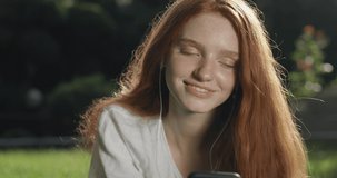 Beautiful girl in headphones with red hair and freckles listens to music using a smartphone. A young woman lies comfortably on the lawn near the house. in slow motion. Shot on Canon 1DX mark2 4K cam