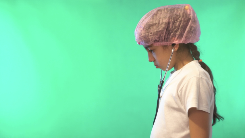A girl in a white t-shirt listens to a Teddy bear. Playing doctor. On the head protection for the hair. Green background. Shooting in the Studio. Medical instrument. 4K video.