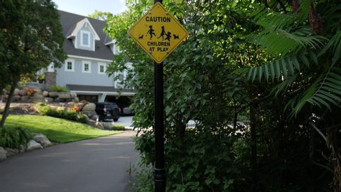 Caution children at play sign on light pole with road to the left and house in the background surrounded by green bushes and trees. Camera pushes into the sign in slow motion.