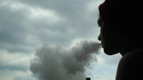 silhouette of young beautiful caucasian woman with a red towel on her head vaping on the balcony after shower. Close up