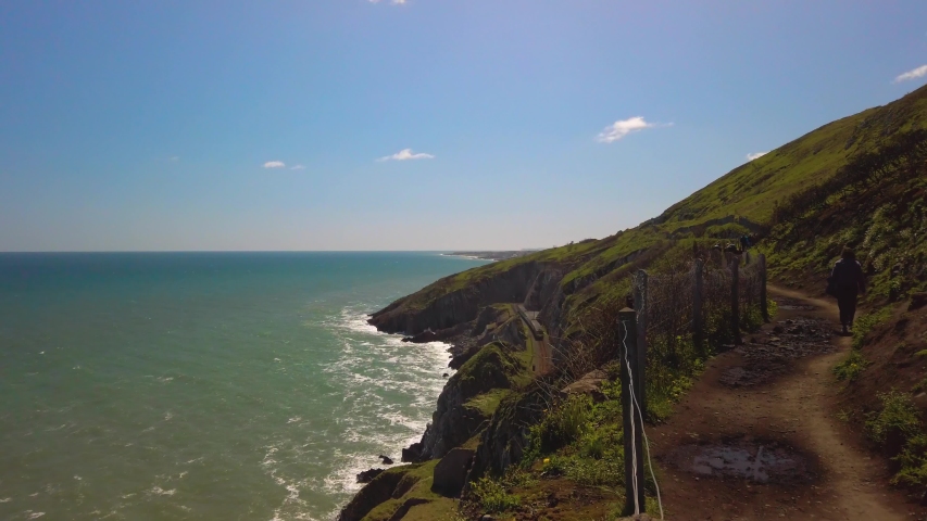 Static clip from a pathway near the coastline of Bray, an Ireland city. Two tourists are walking down the path, while a train in the background is moving on the rails.