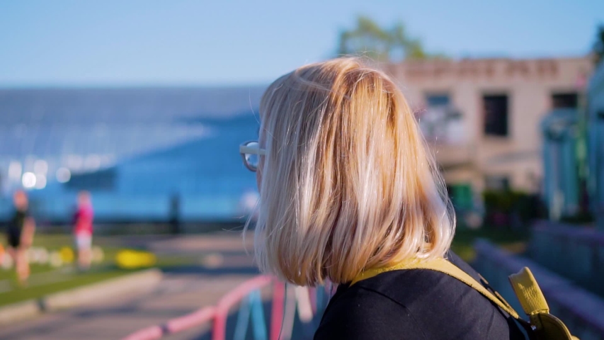 The girl sits on the podium of the school stadium and looks at what is happening on the field. Side view. The girl has white hair, a backpack and headphones in my ears.