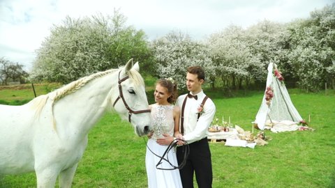 Bride with groom holding white horse and caressing her head to side of them and making wedding teepee making background in nature.