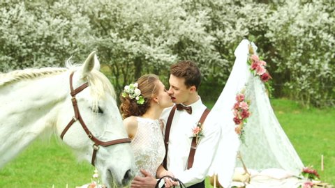 Bride with groom holding white horse and caressing her head to side of them and making wedding teepee making background in nature.