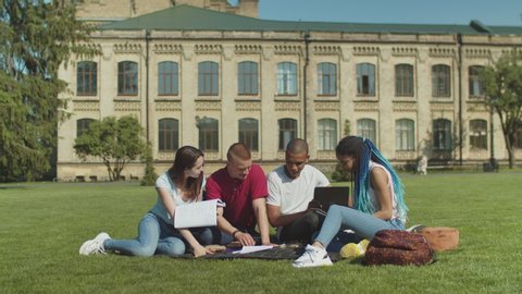 Group of multinational students brainstorming preparing for classes together sitting on grass in front of college. Diverse mates discussing educational material during studying outdoor on campus lawn.