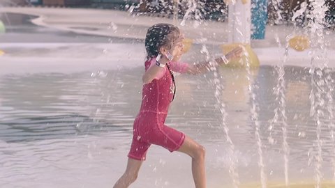 happy adorable girl kid in pink swimsuit runs among high fountain jets at water playground on sunny day slow motion