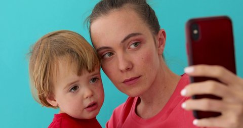 Happy young mother with sad kid taking selfie on mobile phone isolated on blue background