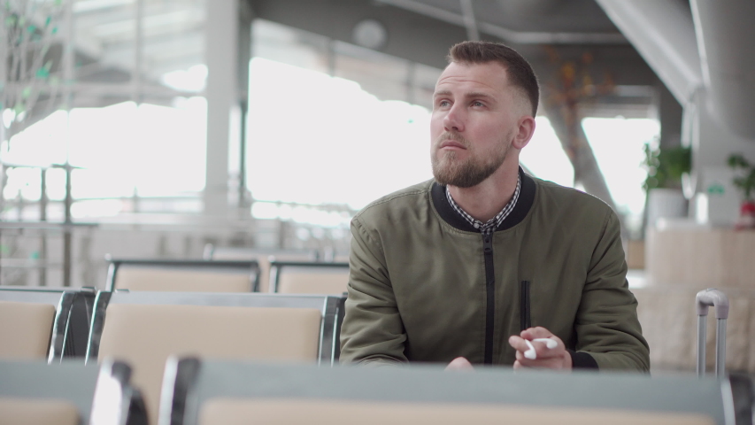 Casual guy in a green jacket looks around in an empty airport and puts his earphone on. Man listening to music on his smartphone while waiting in a terminal.