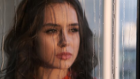 Sad girl looks at the rain outside the window. Beautiful girl looks through a wet window.