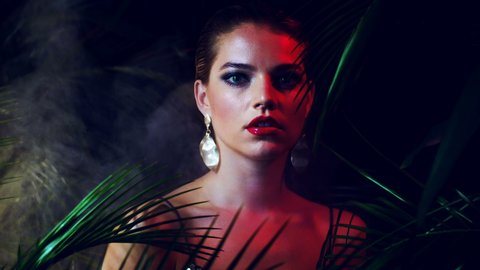 Beautiful girl in luxurious expensive dress, with large stylish gold earrings and ring on her hand, stands among tropical leaves. Concept for advertising jewelry. Red light. Stern look, red lips.