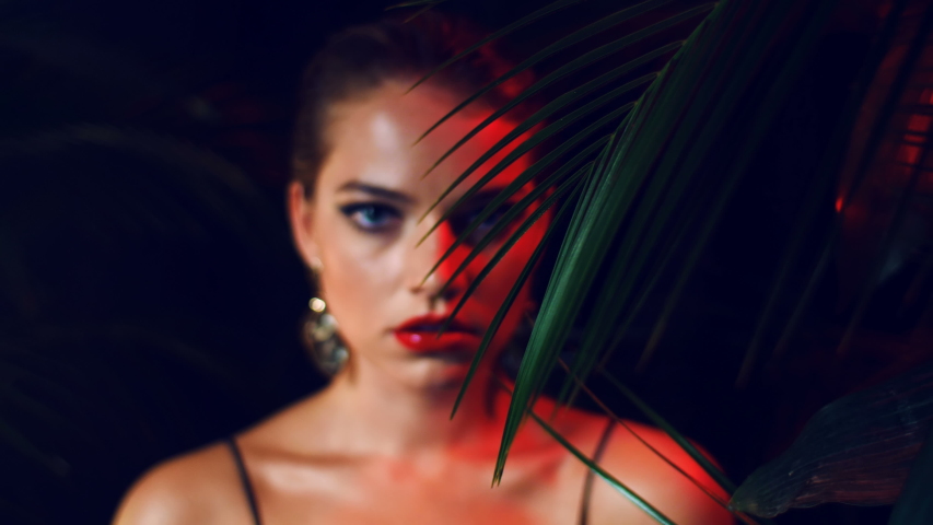 Beautiful girl in luxurious expensive dress, with large stylish gold earrings  stands among tropical leaves.  Concept for advertising jewelry. Red light. Stern look, red lips.