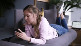 Attractive girl in pink sweater using smart phone while lying on the couch in the living room. Typing, watching video, or browsing social media on smartphone. In front her laptop. Slow motion. Front