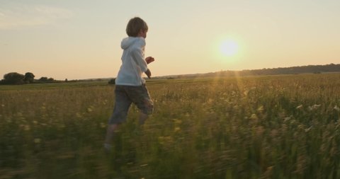 Happy child running on the meadow in the summer while sunset. Happy 7 year old cheerful boy runs and looks back, on field. Happiness concept. Active smiling boy on nature in spring. Slow motion    