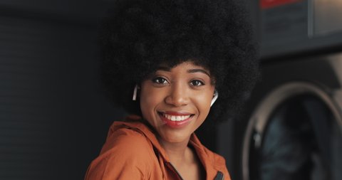 Portrait of young happy African American woman wearing earphones looking into the camera. Self-service public laundry background. Close-up.