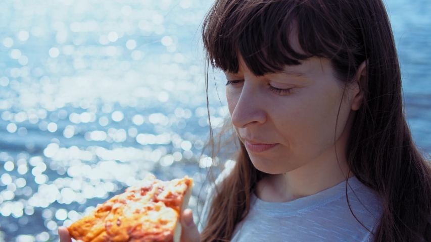 Close-up of young woman bites off piece of pizza and chews it on background of blue sparkling sea.