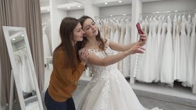 We see 2 brunette girls on video. They are in a wedding salon. One of them is a future bride and another is a consultant in the store. Bride wore her future wedding dress and they making a selfie.