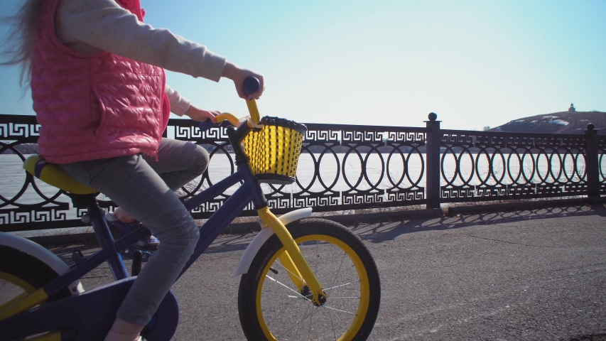 Handheld shot of little blonde girl in protective helmet riding blue first bicycle on an asphalt track with gaps and holes along the embankment of the frozen river. Sport concept outdoors.