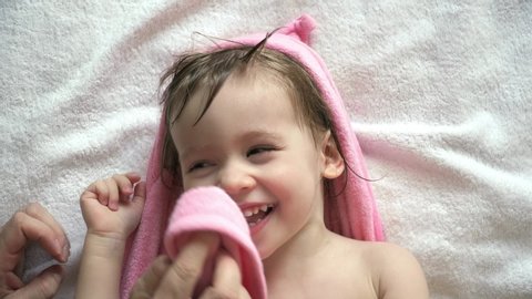 Portrait of little cute caucasian baby girl in pink towel with hood lies on bed after bathing plays and laughs. Father's hands wipe wet face and hair of smiling daughter.