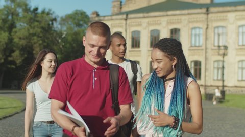 Close-up of cheerful multinational students talking while going through park after classes over college building on background. Happy smiling multi ethnic classmates walking along university campus.