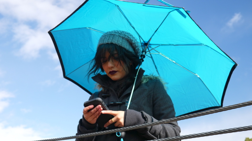 Young woman checking the weather or texting on smartphone with umbrella under blue sky with rain clouds SLOW MOTION.