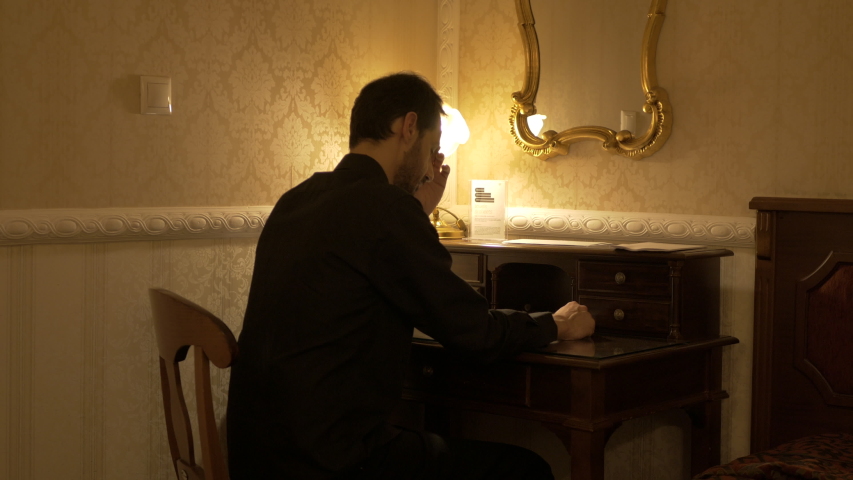 Man wearing black shirt in a dark room with a table light is thinking over a letter that he writes on a wooden desk.