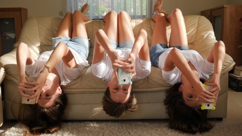 Teenager girls triplet sisters at home watch smart phones in social media lying on couch upside down