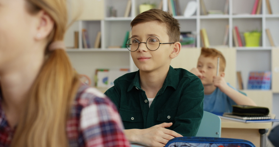 Close up of the cute Caucasian teen small schoolboy in the glasses answering the question of the teacher with rised hand in the lesson.