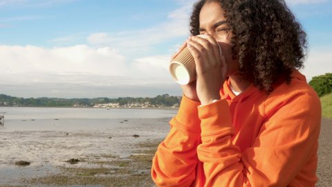 Panning video of beautiful mixed race African American girl teenager young woman wearing orange hoodie and wireless headphones, drinking takeout coffee by a harbor looking sad or thoughtful