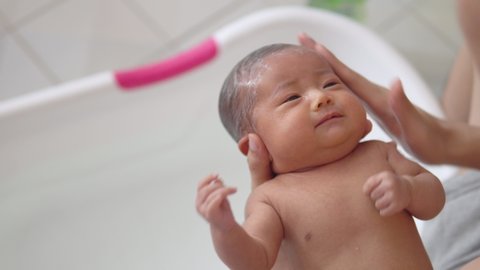 Adorable asian newborn baby girl is washing hair with organic shampoo and taking bath by her father in white plastic tub in bathroom at home. Baby products. Hygiene and health care for newborn baby.
